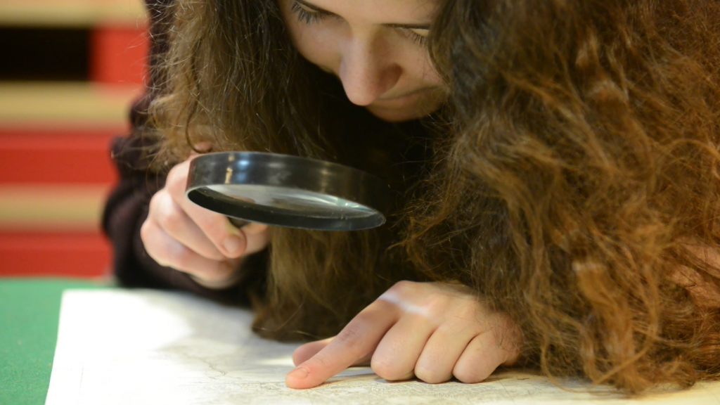 A girl with long hair uses a magnifying glass to inspect an archive document