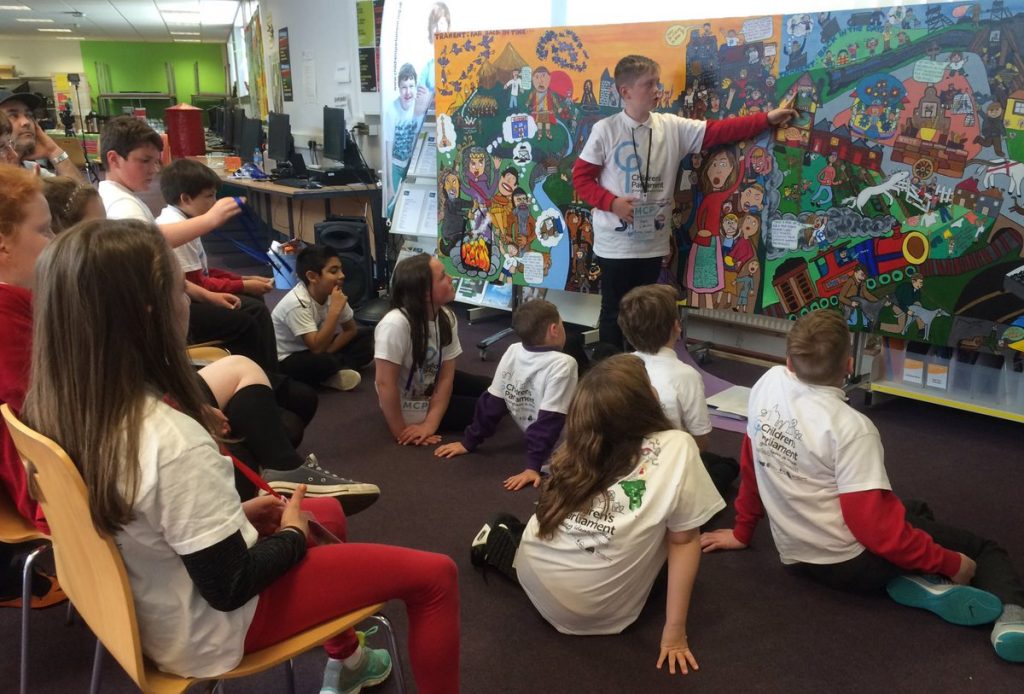 12 children seated and 1 boy standing in front of a mural of Tranent which they are discussing