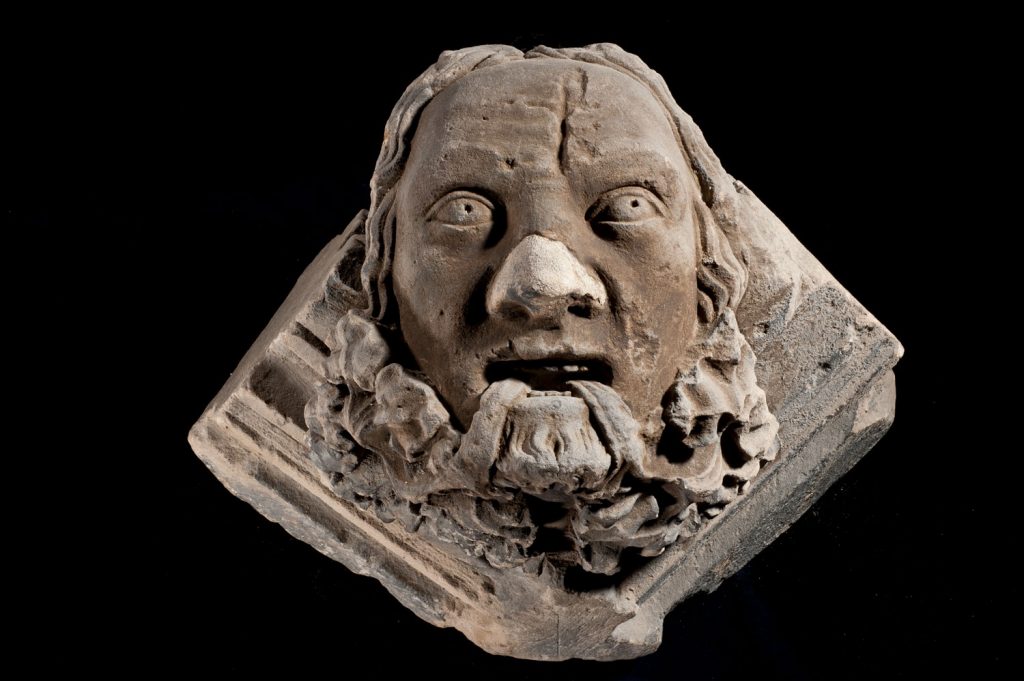 A stone carving of a man's face with thick wavy hair and foliage streaming from his mouth