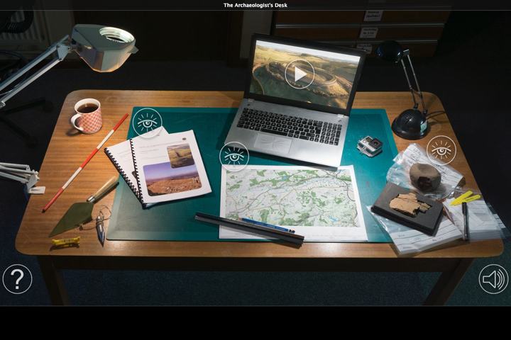 wooden desk with angle poise lamp, trowel, coffee cup, measuring stick, map, research documents, laptop and plastic bags for cataloging archaeological finds 