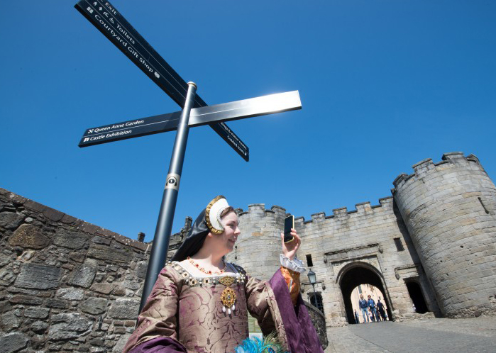 Woman wearing purple and gold corset and white and black headdress holds up a black mobile phone with a grey stone castle entrance behind her and blue sky above