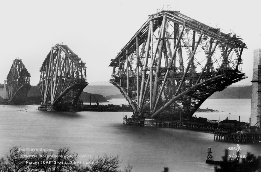 A black and white photograph of a large metal bridge under construction.
