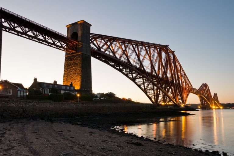 A photograph of a red metal bridge lit up from below, with light reflecting in the water.