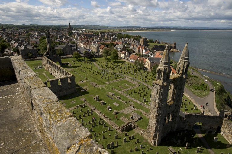 view looking down on a ruined section of a cathedral tower with gravestones and green grass, and in the distance houses and the sea