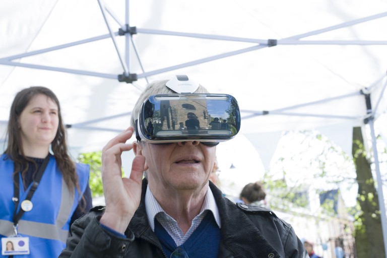 head and shoulders of elderly man wearing a virtual reality headset, looking fascinated by what he can see virtually. A medieval tower is reflected in the surface of the headset