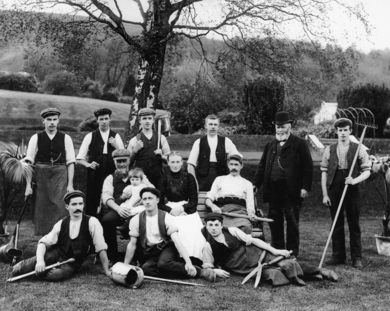 black and white image of a group of men seated and standing in gardens, holding watering cans, shears, rakes and other gardening implements