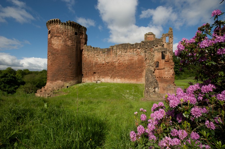 red stone castle with a round tower on a green verge with blue sky above and pink flowers in front