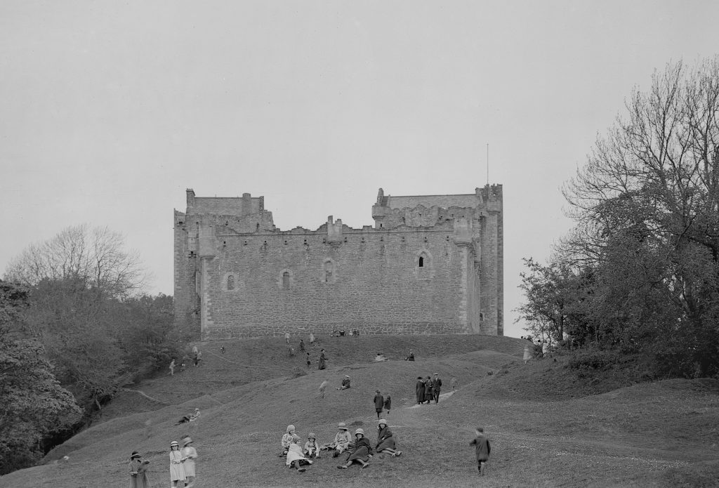 black and white image of castle wall with visitors sitting on verge in front of it 