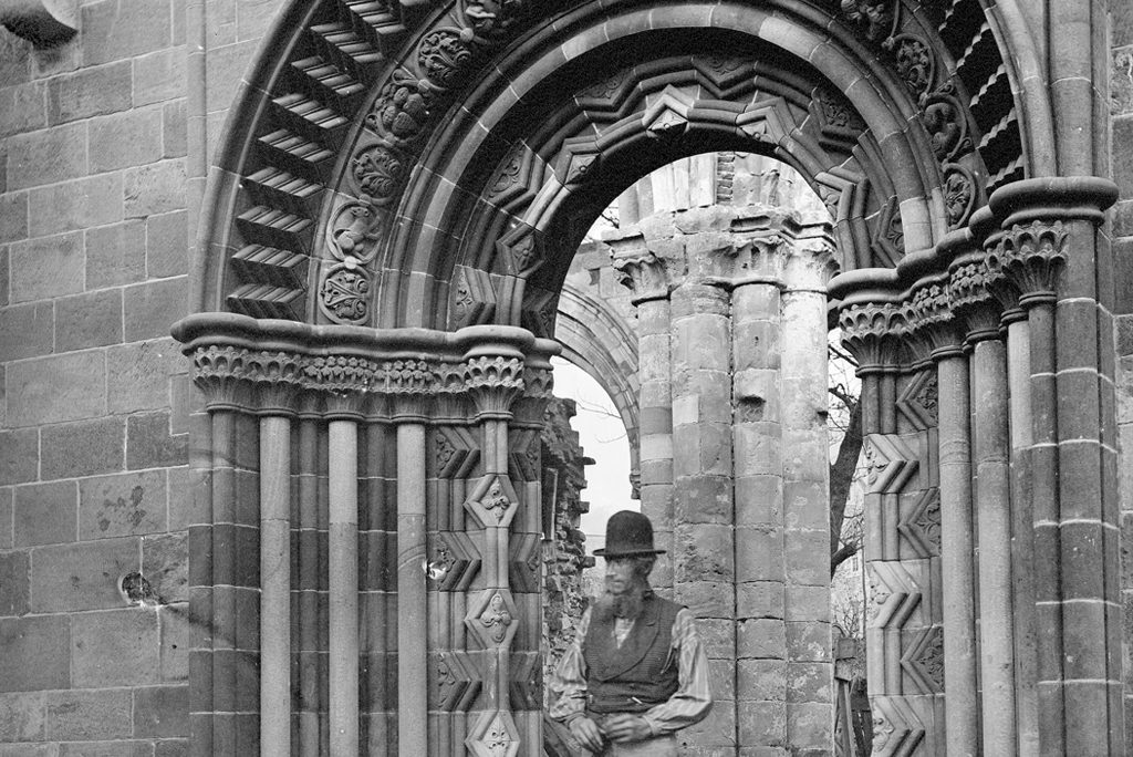 black and white picture of a bearded man wearing a bowler hat and a waistcoat standing under an archway
