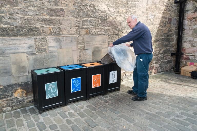 man changes the bag in a bin at the end of a row of bins all labelled in different colours to help people recycle