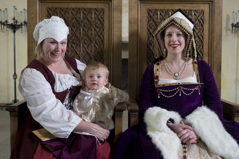 two women in historic costumes sit on wooden thrones, the woman on the left holds a small child with blonde hair, they are smiling for the camera
