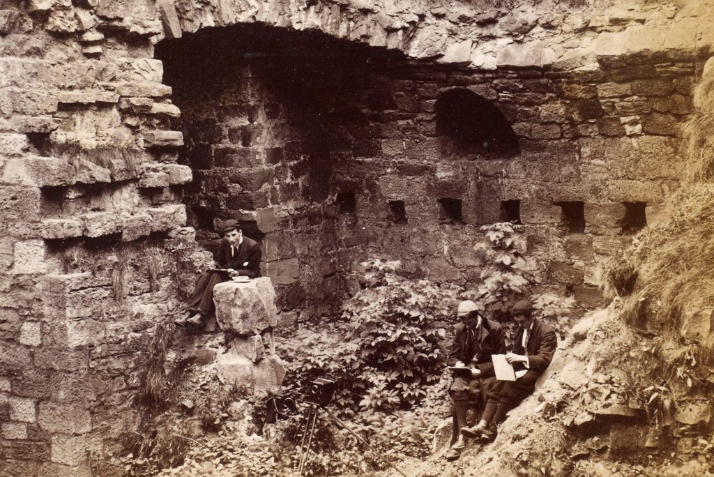 black and white photo of three young visitors in school uniform sitting in the ruins of a historic castle with sketch pads