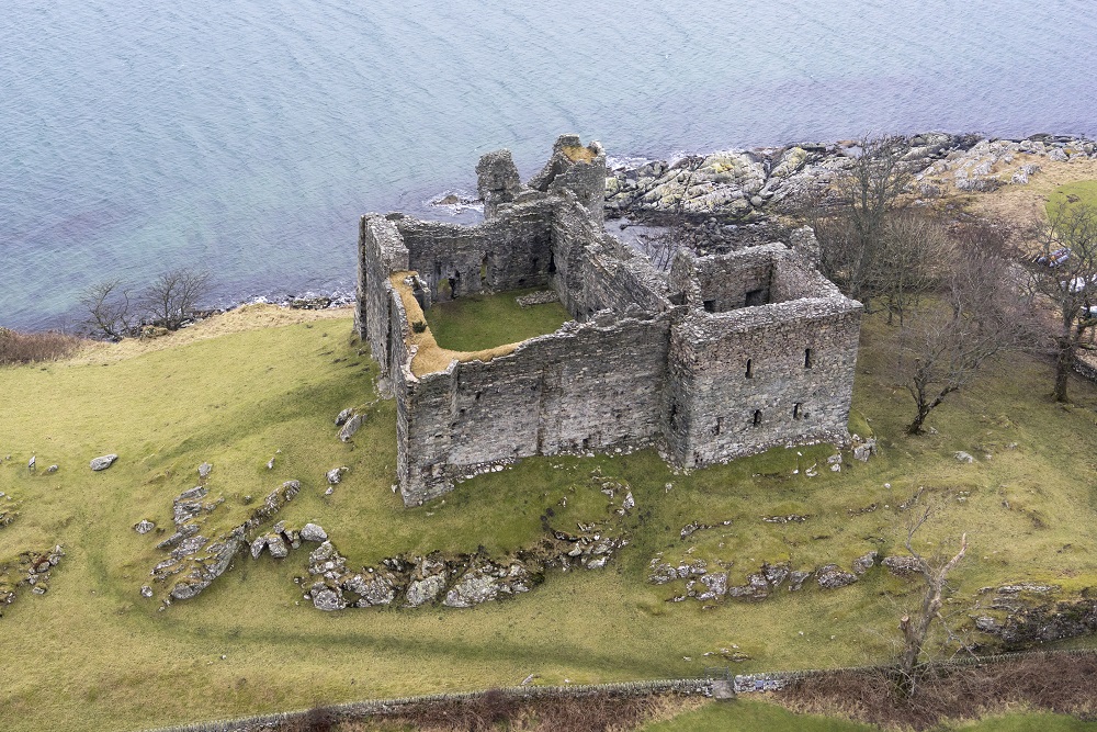 an aerial view of the shell of a grey stone castle on a green hill by a body of water