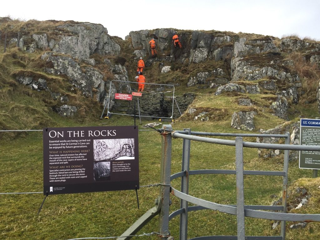 rock face with four figures in orange hi-vis working on a rock face. There is a brown sign in the foreground explaining they are carrying out maintenance to make the rock surface more stable