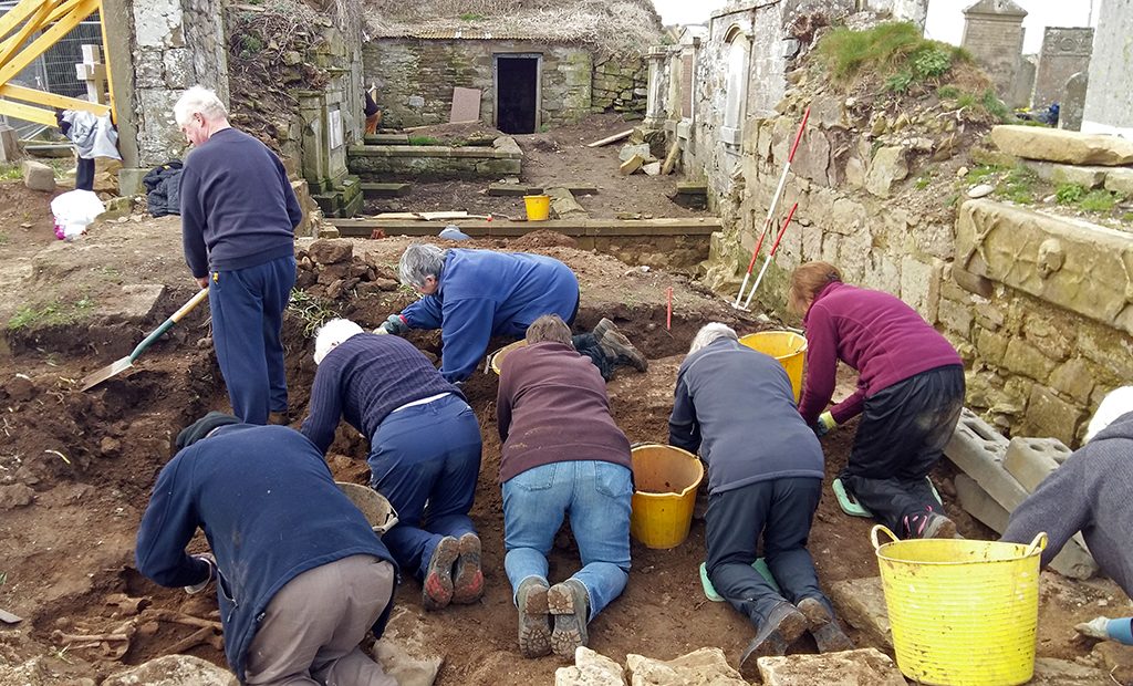 people on their hands and knees in a trench digging up soil to uncover archaeology