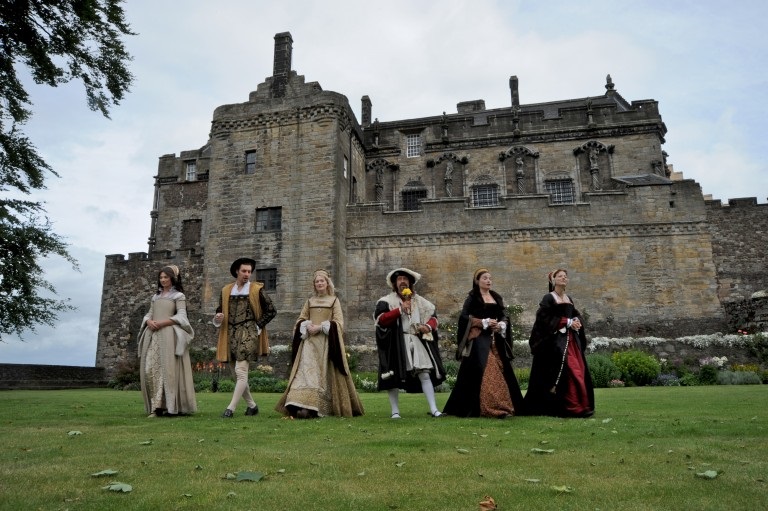 row of people in renaissance costume walking towards the camera with a castle behind them