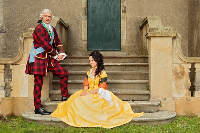 man in tartan suit stands beside woman in yellow gown seated on steps in front of a blue door