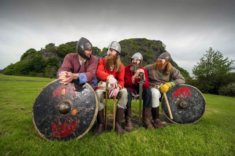 four men in viking outfits sit in a row talking to each other. They are holding shields and swords