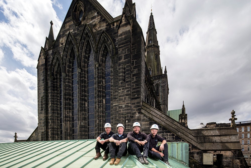 Four men sitting in a row on a green surface dressed in black and wearing hard hats with a cathedral behind them