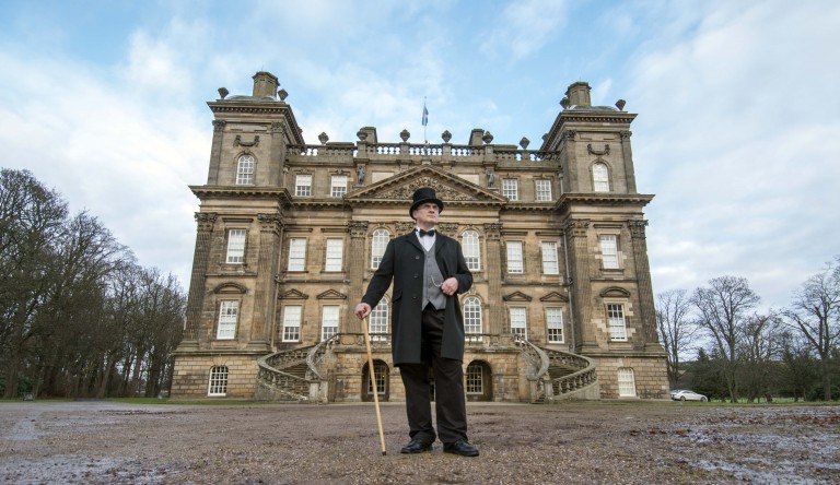 man in victorian dress stands leaning on a cane with a mansion in the background behind him