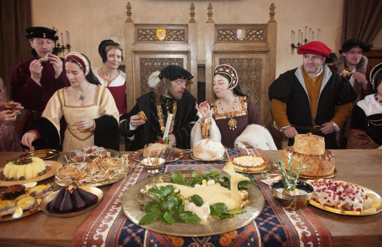 a table laid out with old fashioned types of food, with a row of people in renaissance costume behind including two royals seated on wooden thrones