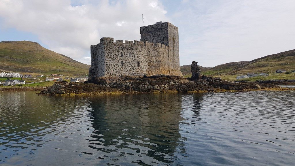 a grey stone castle with hills in the distance behind it and a body of water in front of it
