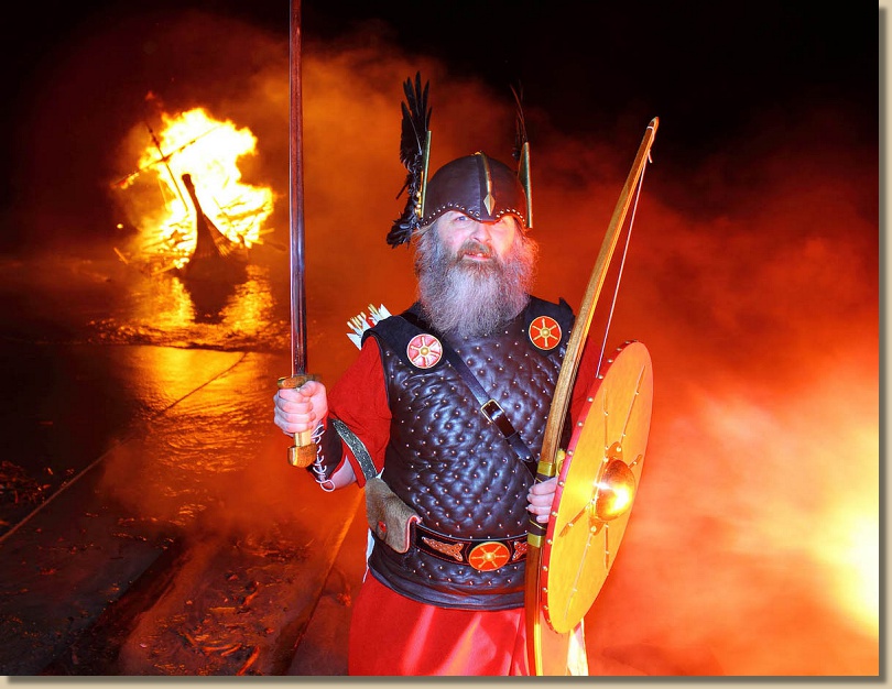 bearded man in helmet and armour stands before a burning boat