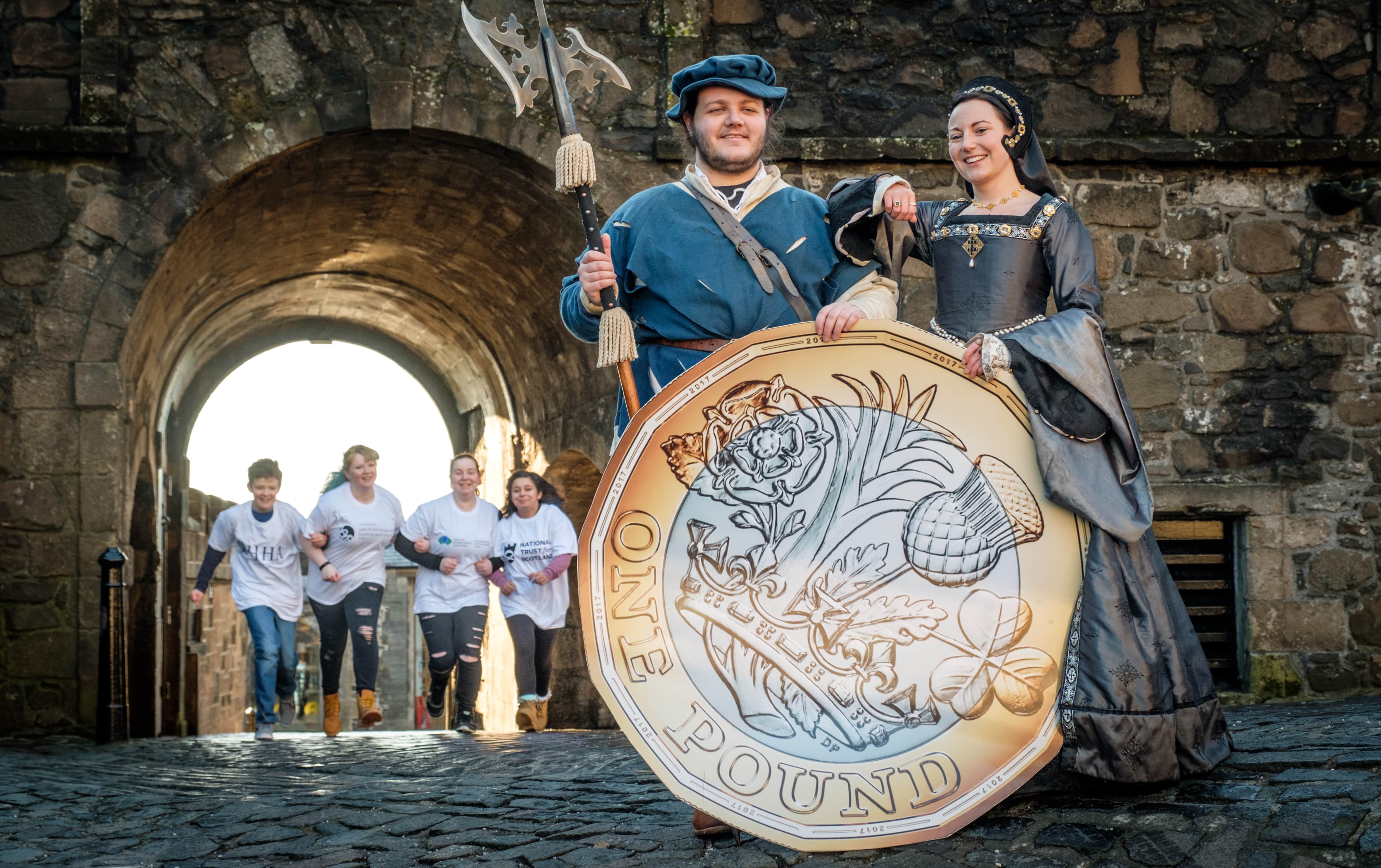 two people in historic costumes hold a giant pound coin whilst behind them a row of four young people run towards them