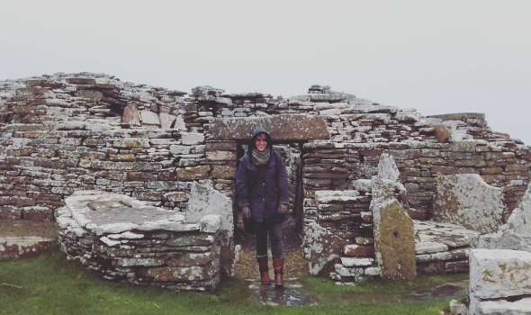 person bundled up in rain coat, boots and gloves smiles at the camera as she stands before an ancient stone building