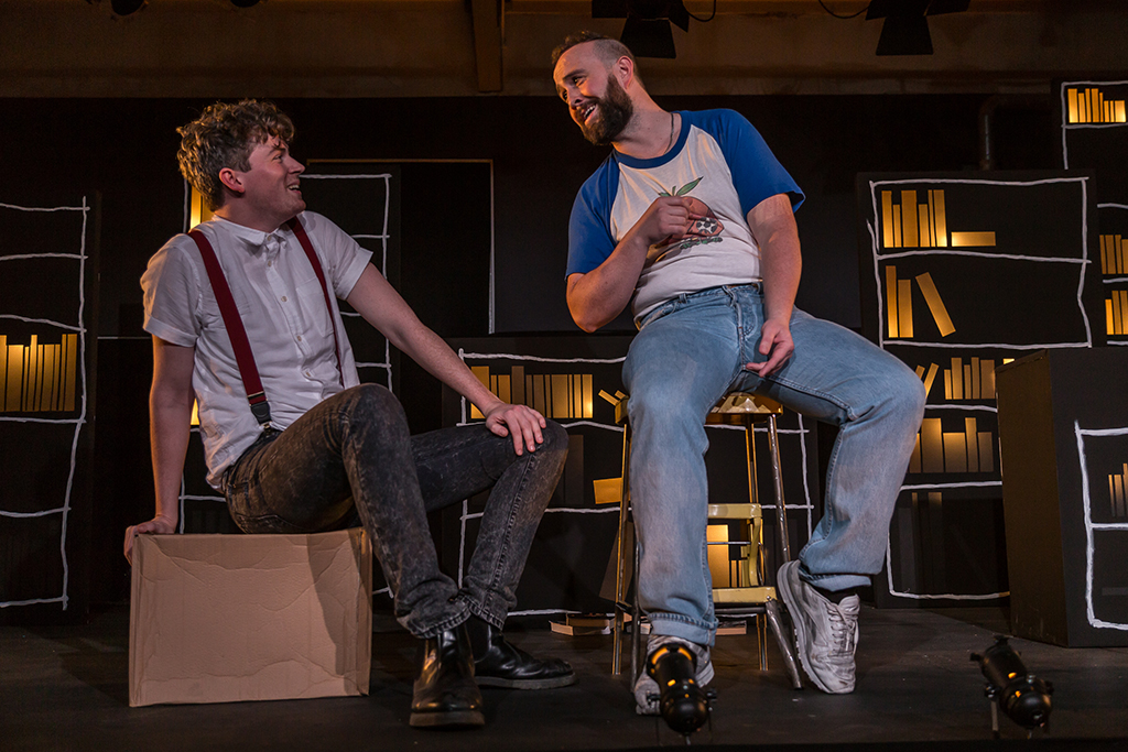 two men sit on stage talking to each other, smiling
