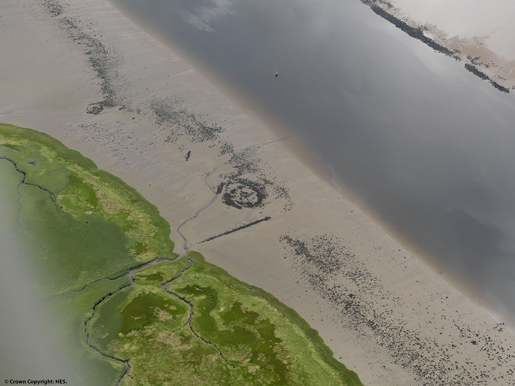 view from above of sandy beach with grey circle in the middle, lined at either side by green land and grey water