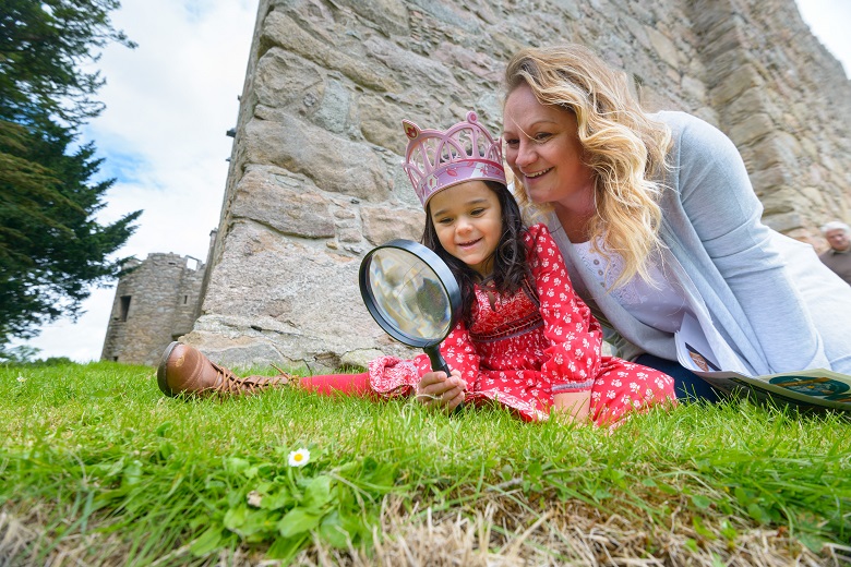 Mother and daughter sit at the foot of a castle and explore through a magnifying glass
