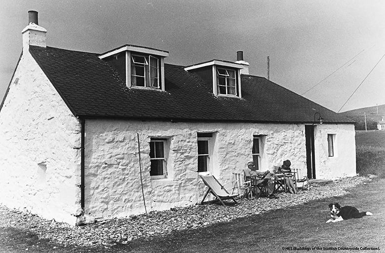 1970s, a couple sits outside their cottage on deck chairs with a collie dog lying in the road.