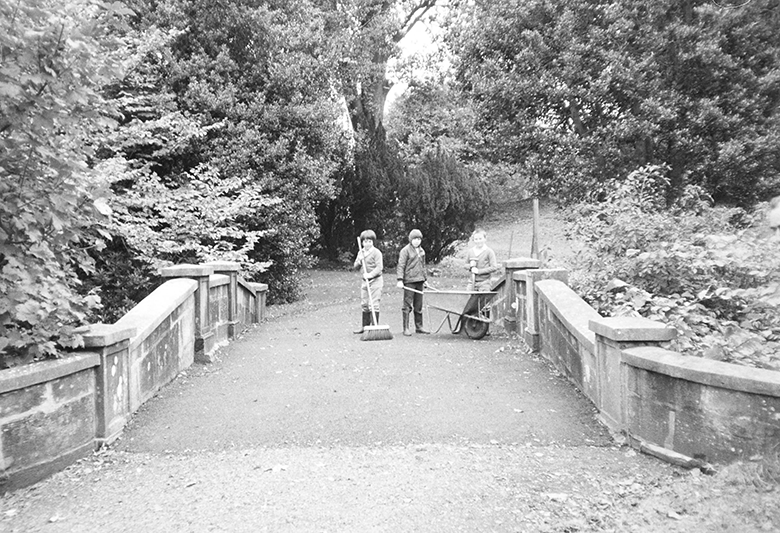 Three children stand on a bridge with a wheelbarrow and brush. 1970s Scotland.