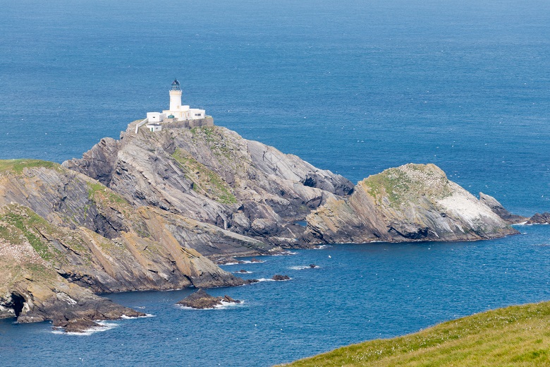 View of Muckle Flugga island. A white lighthouse sits atop a rocky outcrop in a calm blue sea