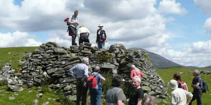 a group of people with backs to the camera congregate around remains of a stone building