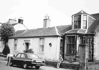 Black and white photo of a residential street in the 1970s. A boy sits on the garden wall while his dad fixes the car.