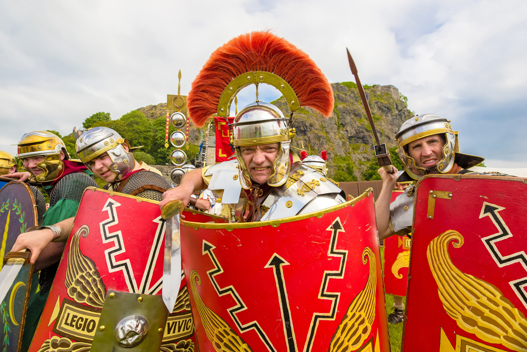 four men in roman helmets charge towards camera holding weapons and shields
