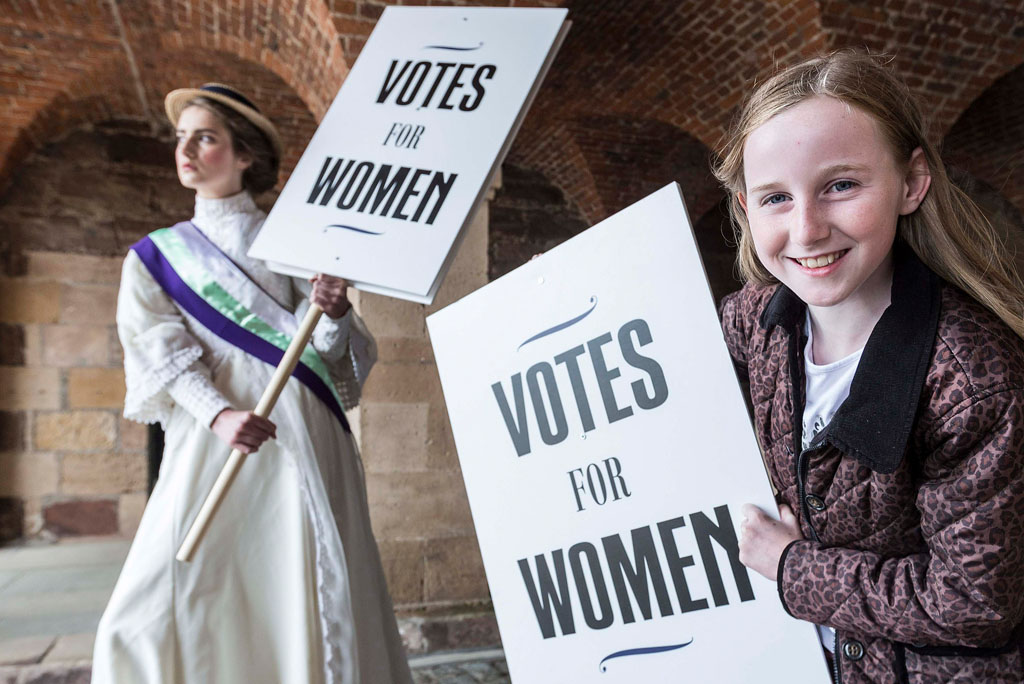 woman in victorian dress and young girl in modern dress hold signs saying 'votes for women'