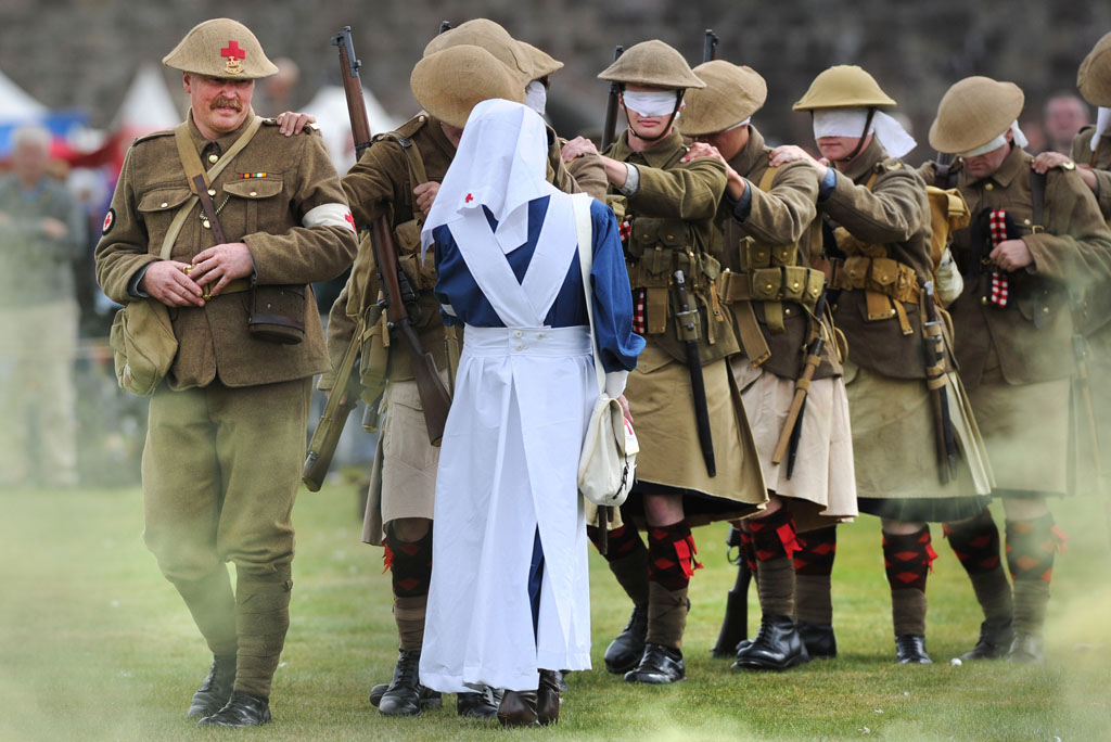 queue of seven men wearing military uniform and blindfolds walk in single file across a field as a nurse goes to meet them