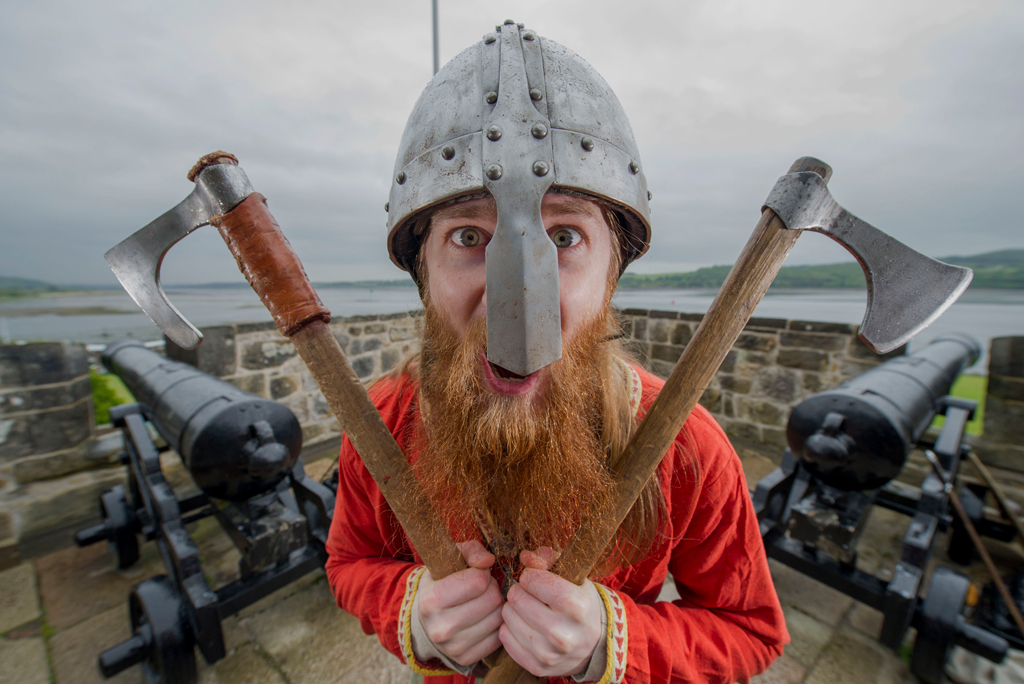 man with helmet covering nose holds two axes and bellows at camera