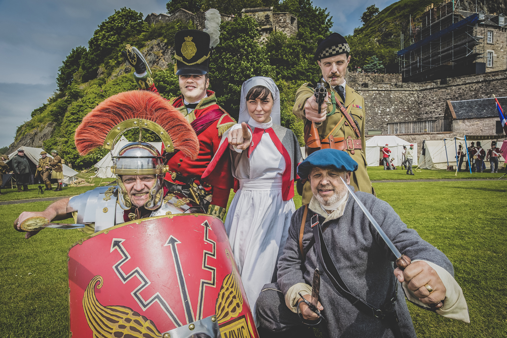 five people in different historic costumes point at camera with castle behind them