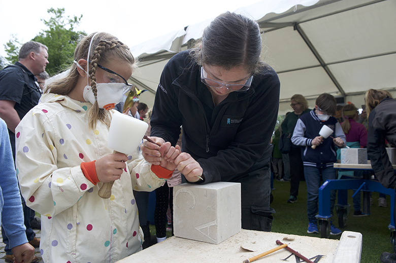 Young girl has a go at carving a stone block, supervised by an adult