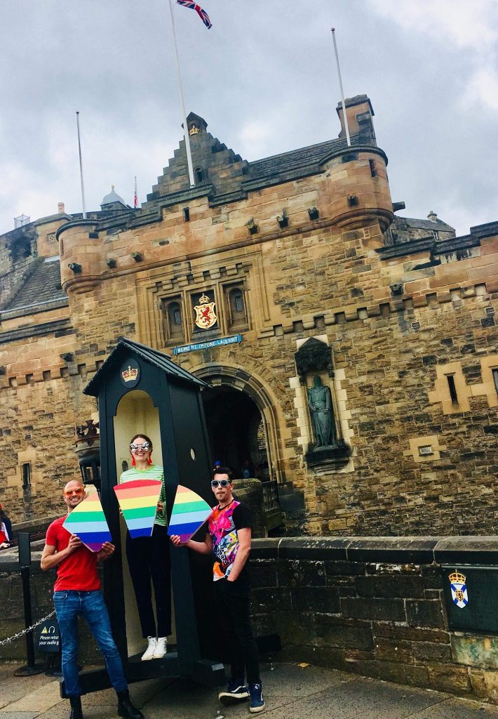A photograph of three people dressed in colourful clothes outside of a castle, holding a keystone in rainbow colours.
