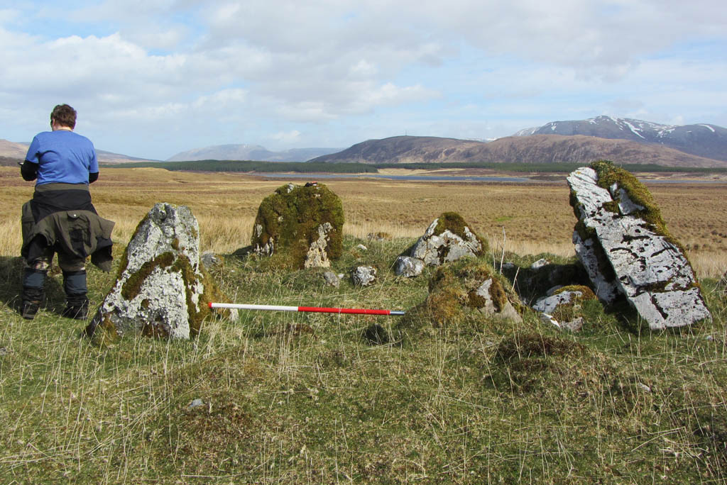 person with back to camera standing beside a stone circle with mountains in distance