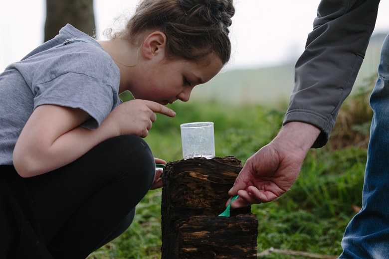 A young girl squats on the ground peering into a cup that might have insects or other small wildlife in it