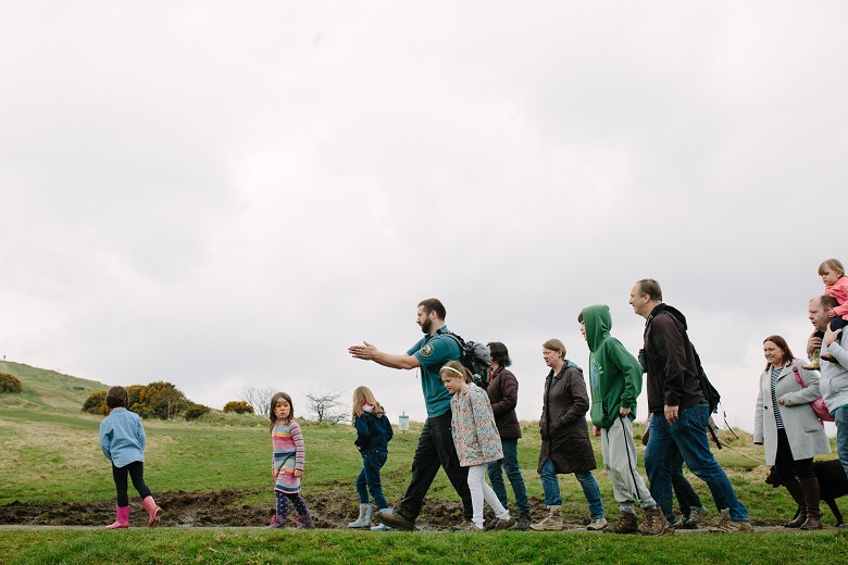 A ranger leads a tour group through a park landscape. The group includes young children, adults and older people
