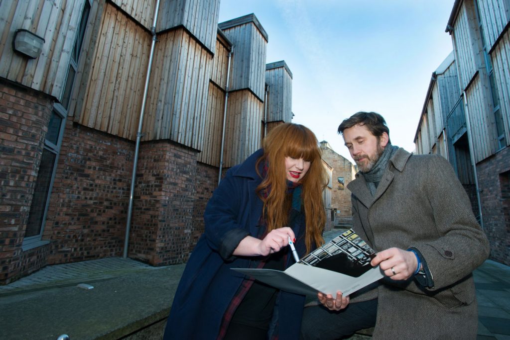 A woman with long ginger hair and red lipstick with a man in a woolly coat standing beside a brick and wooden building, looking at a document