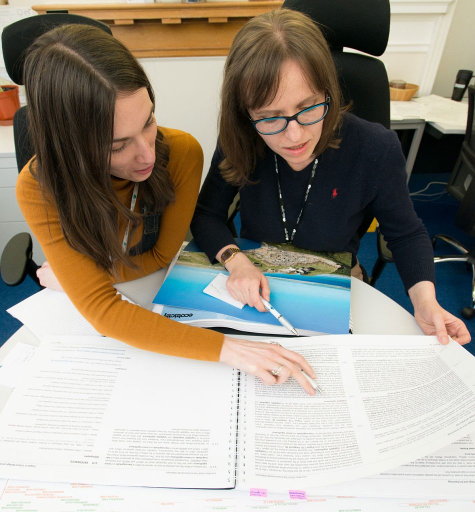 Two women with long brown hair talking and looking at paperwork on a table