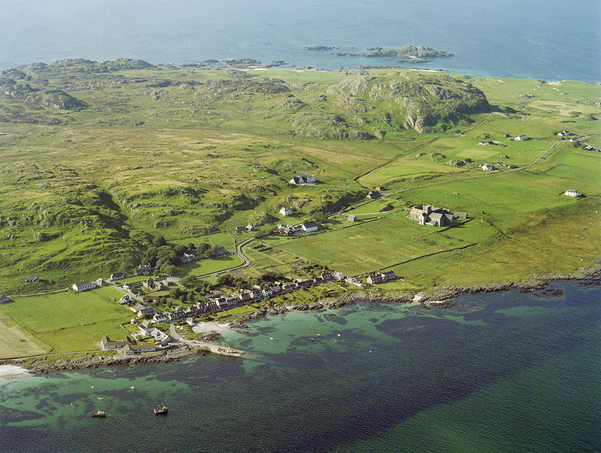 An aerial view of the island of Iona. The island is green and dominated by rocky hills. A row of houses stands in front of a small bay and jetty. Iona Abbey is located nearby,